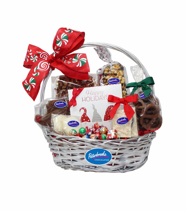 A wicker gift basket with a red and white holiday bow, filled with an assortment of chocolate covered treats including popcorn, caramel corn, assorted chocolates, pretzels, peppermint bark, and cookies.