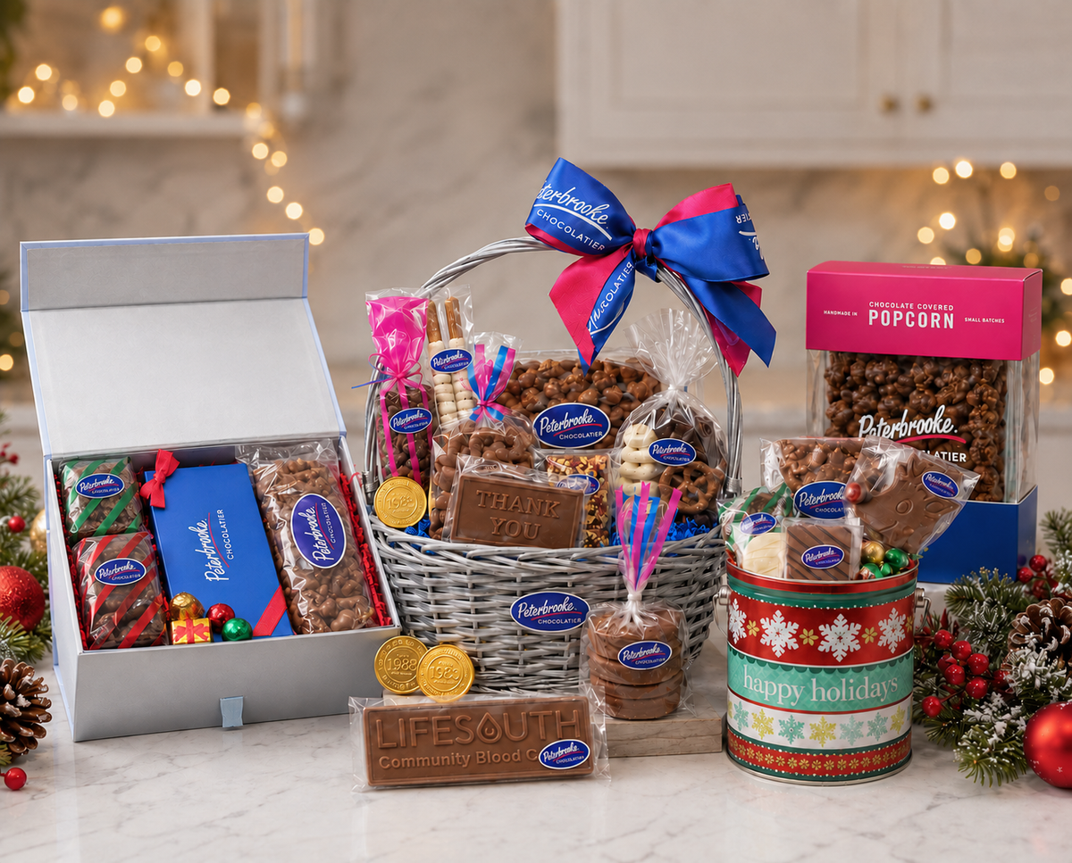 A festive holiday gift basket filled with assorted Lifesouth chocolates, cookies, and treats, displayed alongside an open gift box and a box of popcorn on a marble countertop with holiday decorations and warm string lights in the background.