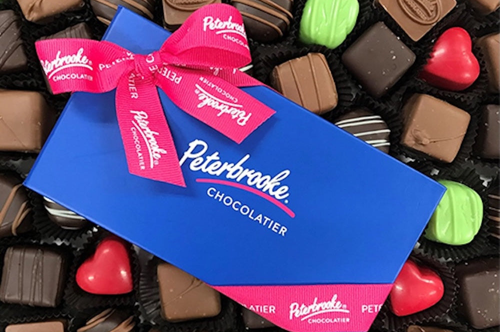 Blue box of Peterbrooke Chocolatier chocolates with a pink ribbon, surrounded by assorted chocolates