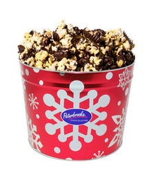 A red metal tin with white snowflake designs filled with dark chocolate covered popcorn.